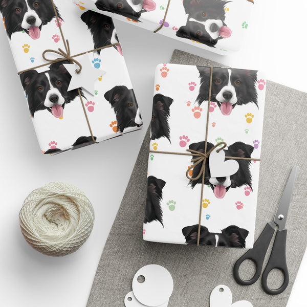 Three gift boxes wrapped in a white paper featuring a pattern of black and white Border Collie dog faces and colorful paw prints, tied with twine and decorated with blank white tags. The scene is styled with a spool of twine, scissors, and paper circles on a gray and white surface.