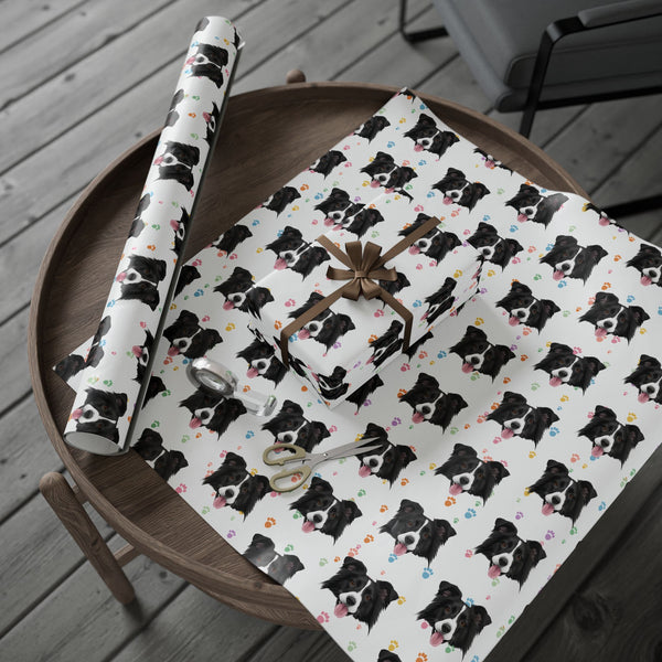 White wrapping paper with a repeating black and white dog print and small colorful heart accents, shown wrapped around a gift box and a matching rolled sheet on a round wooden table. A gift bag, ribbon, and scissors are also visible.