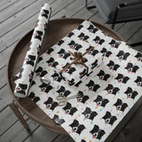 White wrapping paper with a repeating black and white dog print and small colorful heart accents, shown wrapped around a gift box and a matching rolled sheet on a round wooden table. A gift bag, ribbon, and scissors are also visible.