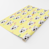A roll of wrapping paper with a repeating pattern of white dog faces on a pale yellow background, accented with small pastel paw prints, shown on a white studio background.