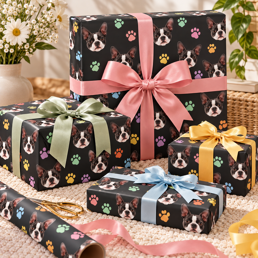 Four wrapped gift boxes arranged on a cream woven rug, all featuring black wrapping paper printed with Boston Terrier dog faces and multicolored paw prints. The boxes are tied with satin ribbons in pink, sage green, yellow, light blue, and dark blue, with a roll of matching wrapping paper and gold scissors in the foreground.