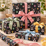 Four wrapped gift boxes arranged on a cream woven rug, all featuring black wrapping paper printed with Boston Terrier dog faces and multicolored paw prints. The boxes are tied with satin ribbons in pink, sage green, yellow, light blue, and dark blue, with a roll of matching wrapping paper and gold scissors in the foreground.