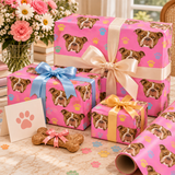Three gift boxes wrapped in bright pink dog-face patterned paper, tied with cream, blue, and yellow ribbons, arranged on a marble-like surface with a rolled matching wrapping paper, a paw print card, and a bone-shaped treat.