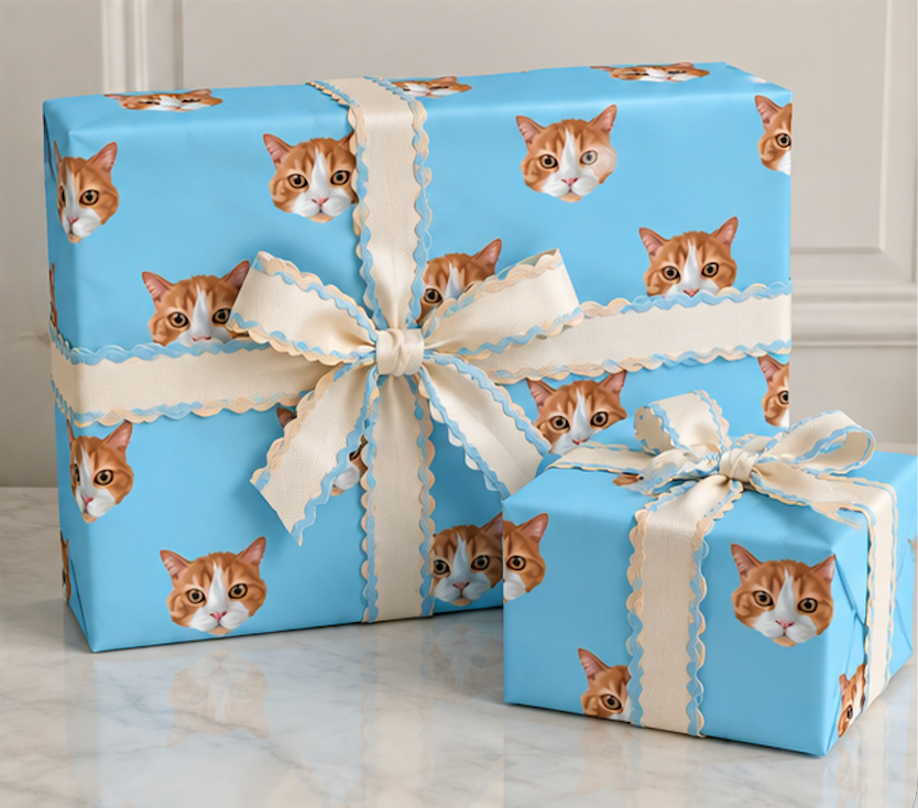 Three wrapped gift boxes on a light marble surface against a white paneled wall. The boxes are wrapped in bright blue paper printed with repeated orange-and-white cat faces, and each is tied with a cream ribbon featuring a scalloped edge and a large bow.