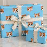 Three wrapped gift boxes on a light marble surface against a white paneled wall. The boxes are wrapped in bright blue paper printed with repeated orange-and-white cat faces, and each is tied with a cream ribbon featuring a scalloped edge and a large bow.