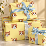 Three wrapped gift boxes stacked on a light marble surface, covered in pale yellow wrapping paper printed with orange-and-white cat faces and tied with blue-and-white gingham ribbons. A vase of white daisies sits to the left, and a cream wall is in the background.