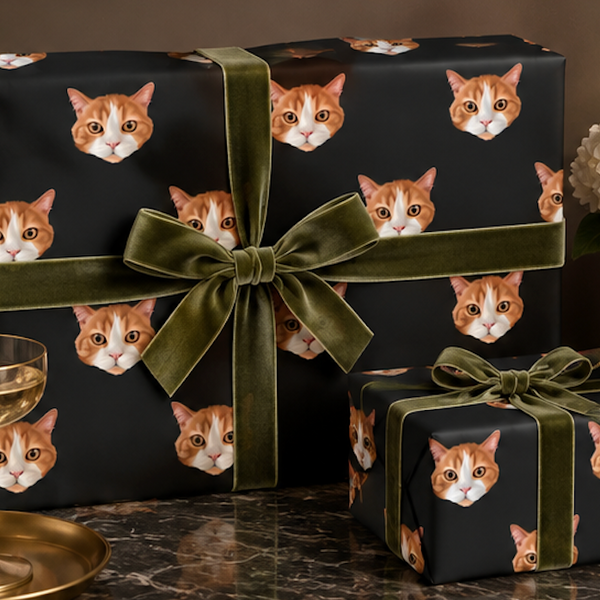 Two gift boxes wrapped in black paper printed with repeated orange tabby cat faces, tied with olive green velvet ribbon and bow, styled on a dark marble surface with a glass and white flowers in the background.