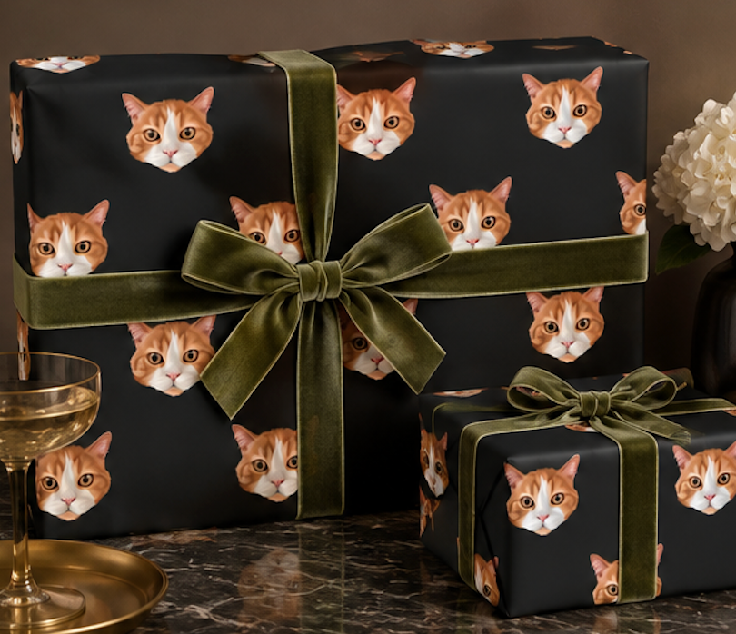 Two gift boxes wrapped in black paper printed with repeated orange tabby cat faces, tied with olive green velvet ribbon and bow, styled on a dark marble surface with a glass and white flowers in the background.