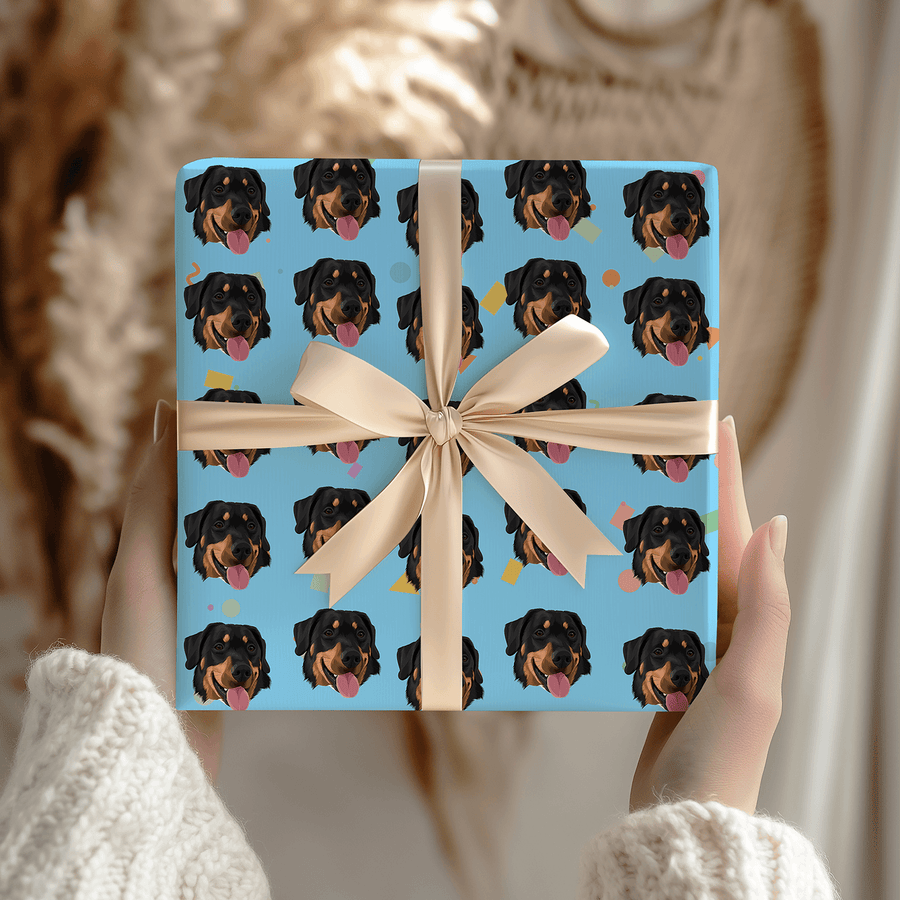 A square gift box wrapped in light blue paper printed with a repeating pattern of Rottweiler dog faces, tied with a cream ribbon and bow, held by hands in a cozy indoor setting.