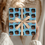 A square gift box wrapped in light blue paper printed with a repeating pattern of Rottweiler dog faces, tied with a cream ribbon and bow, held by hands in a cozy indoor setting.